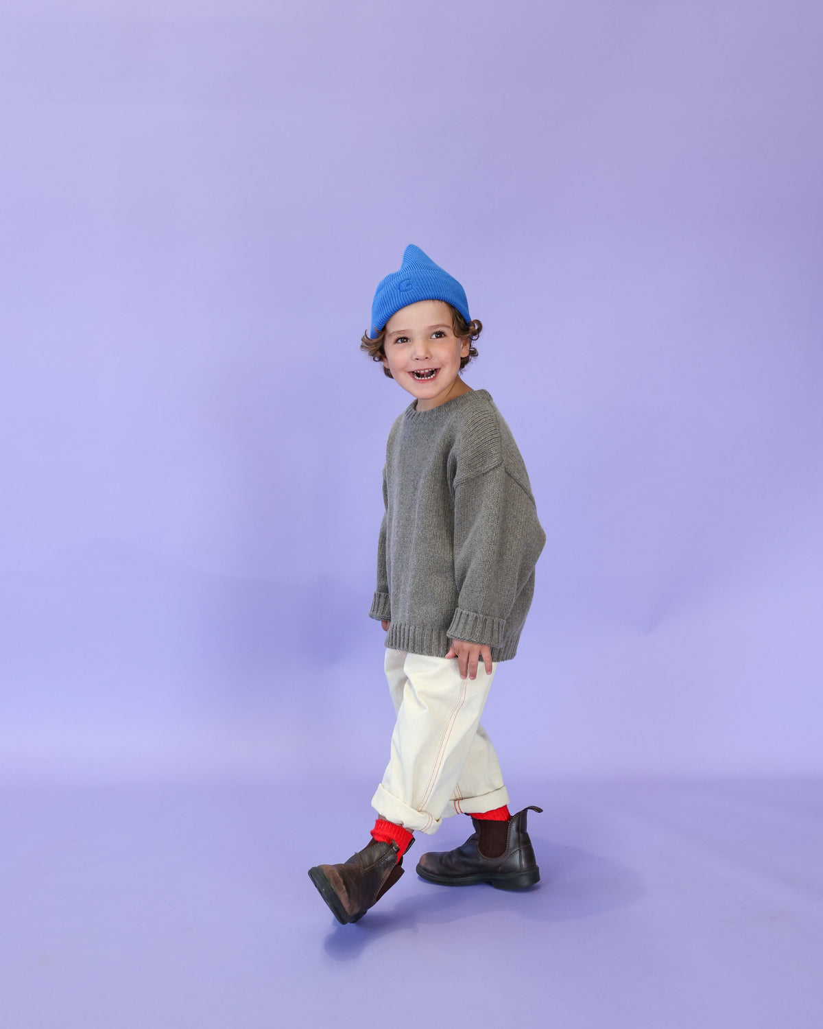 A child wearing a gray pullover with rib finish at neck, hem and cuff, featuring a relaxed fit and a dropped shoulder. The child is also wearing a blue beanie, white pants, and black boots with red socks.