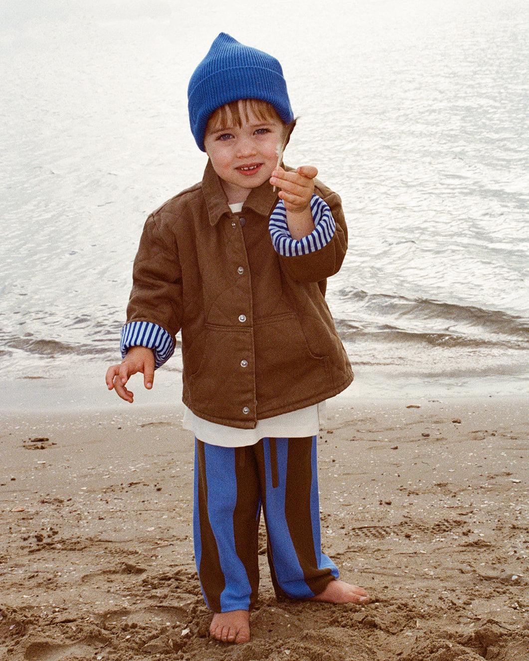 A child standing on a beach wearing a brown organic quilted denim jacket with a blue striped collar and blue pants. The child has a blue beanie on and is holding a stick.