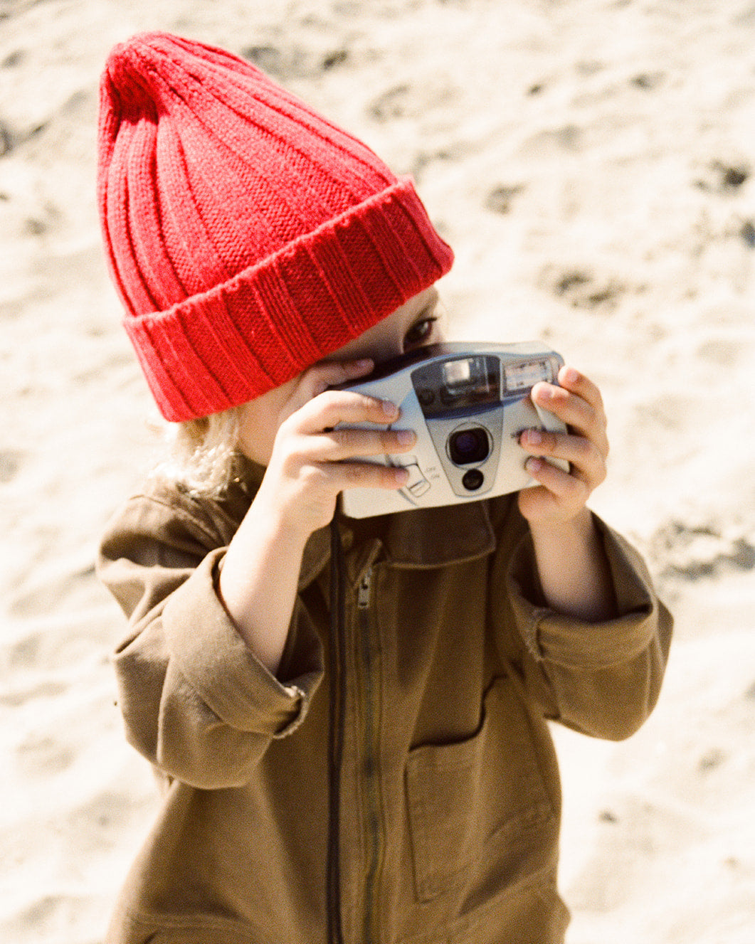 Person wearing a red beanie and brown coat holding a vintage camera on a sandy background
