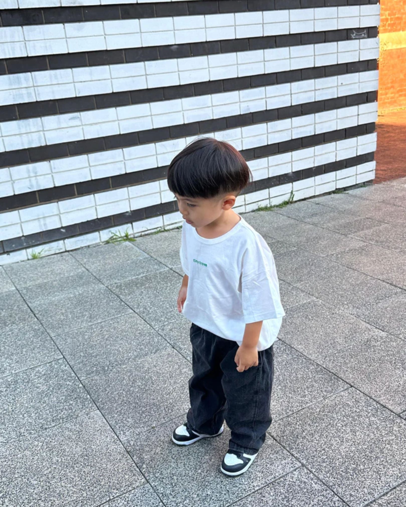 A child standing on a tiled floor against a wall, wearing a white t-shirt and black denim jeans with a pair of white sneakers.