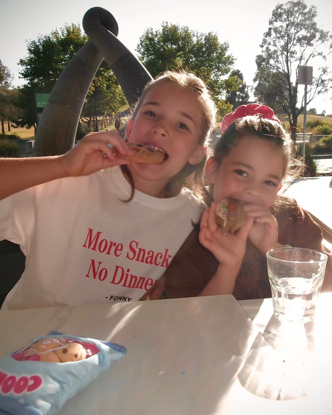 Two children sitting at a table outdoors, wearing white t-shirts with the text 'More Snack No Dinner' on them, and eating snacks.