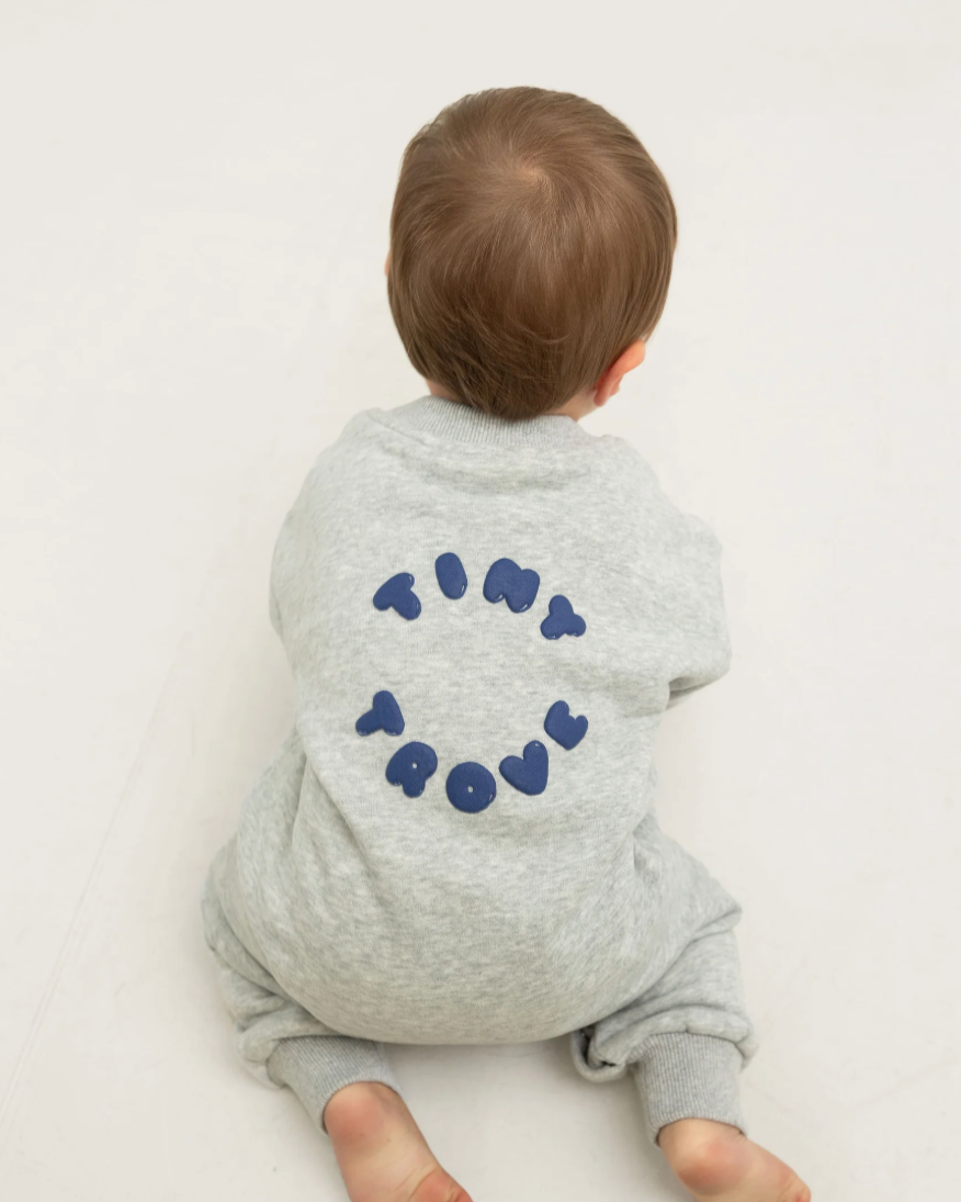 Baby wearing a grey onesie with 'TINY TROVE' printed on the back against a white background