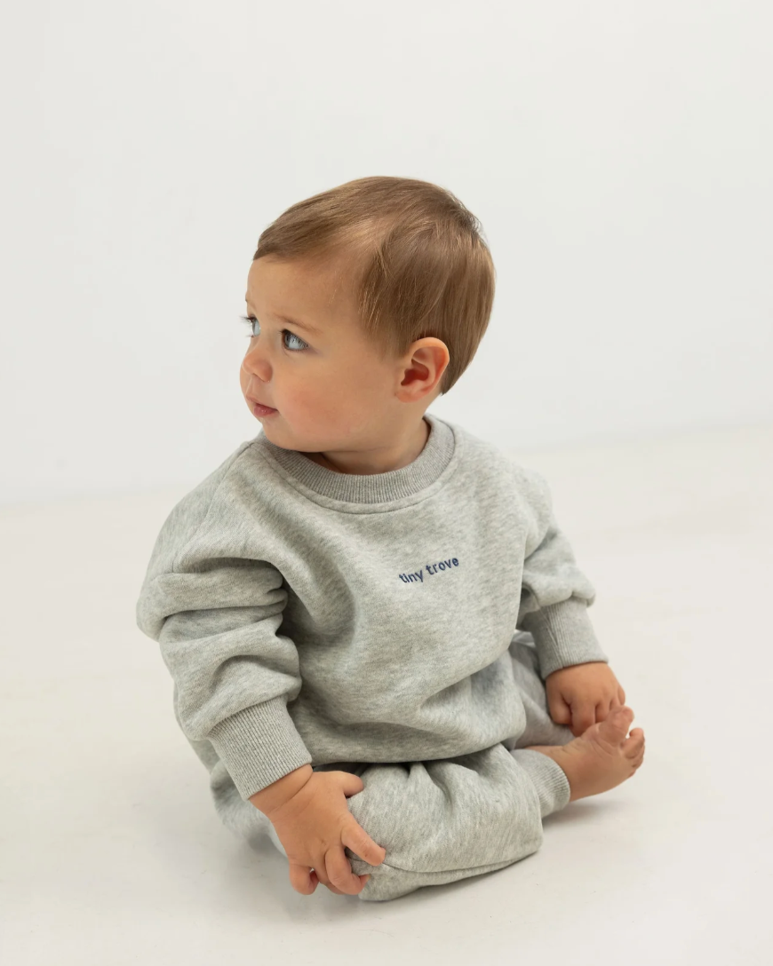 A baby sitting on a white surface wearing a gray marled romper with 3D logo detailing on the front and a puff circle logo on the back.