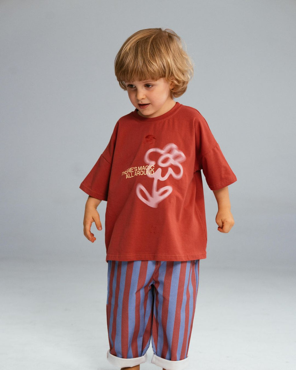 A child wearing an oversized burgundy tee with a contrasting white flower and text design, paired with striped pants.